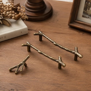 May include: Two long cabinet pulls and one smaller cabinet knob, all crafted from metal with a textured, antique brass finish. Each piece is shaped like a natural tree branch with realistic knots and irregular, organic twig-like extensions.