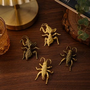 May include: Four metallic gold-toned insect figurines arranged on a surface. Two of the figures are shaped like mantises with textured wings and legs, while the other two are shaped like crickets with detailed segmented bodies and antennae.