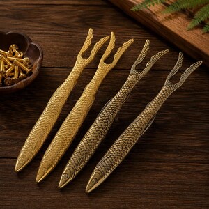 May include: Four metallic fish-shaped forks with detailed scale textures and bifurcated tails. The forks have a golden finish, with two appearing brighter and two having a darker, antique-style patina. Each fork features a pointed head.