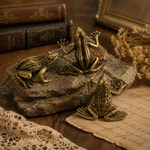 Puede incluir: Tres pequeñas figuras de ranas de latón de estilo antiguo con piel texturizada y detallada. Una rana está colocada de perfil, mientras que las otras dos se muestran desde una perspectiva cenital, todas con un acabado dorado metálico y rasgos anatómicos realistas.