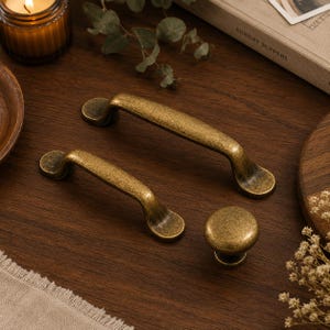 May include: Two brass-toned metal drawer pulls with a distressed, antique finish and one matching round cabinet knob. The hardware features curved, ergonomic shapes with rounded ends and a textured, weathered surface appearance.