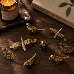 May include: A collection of seven brass-toned metal decorative objects including two birds in flight, two perched birds, two fan-shaped pieces, and one small fluted cylindrical knob. The items are arranged on a dark wooden surface near a candle.
