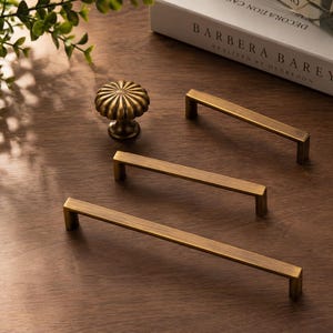 May include: A collection of antique brass hardware including one round, fluted cabinet knob and three rectangular, linear cabinet pulls of varying lengths, all featuring a brushed metallic finish and a simple, clean-lined design.