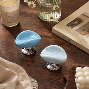 May include: Two cabinet knobs with smooth, rounded, light blue ceramic tops and polished silver-tone metal bases. The knobs feature a concave, pebble-like shape with a glossy finish, designed for attachment to furniture drawers or cabinet doors.
