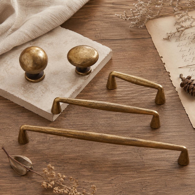 May include: A collection of antique brass hardware including two round cabinet knobs and three elongated drawer pulls of varying lengths. Each piece features a textured, weathered metallic finish with a warm, muted gold tone and tapered bases.