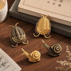 May include: Two metallic trilobite figurines and two metallic snail figurines. One trilobite is finished in a dark antique bronze tone, while the other is a bright gold tone. Both snails feature textured bodies and smooth spiral shells.