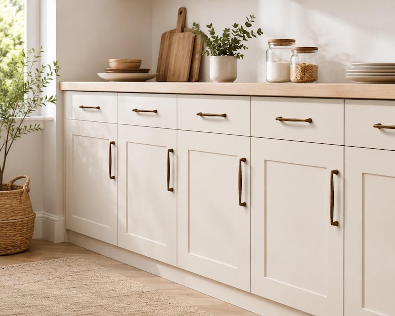 May include: Off-white kitchen cabinets featuring shaker-style doors and drawers with dark bronze metal hardware pulls. The cabinets are topped with a light wood countertop, creating a clean and functional storage space for a kitchen area.