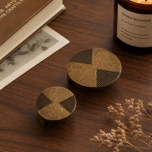 May include: Two circular cabinet knobs with a brushed antique brass finish. Each knob features a geometric pattern of alternating smooth surfaces and recessed diagonal lines arranged in a cross-like design across the round face of the hardware.