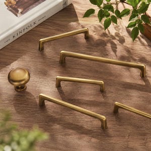 May include: A collection of brushed brass cabinet hardware including four rectangular bar pulls of varying lengths and one round mushroom-shaped knob. Each piece features a smooth, metallic finish with clean lines and a consistent golden tone.