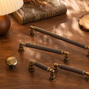 May include: A collection of four cabinet hardware pieces including one round knob and three bar pulls. Each piece features a dark, textured, knurled metal center with antique brass-toned end caps and mounting bases for a cohesive hardware set.