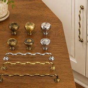 May include: A collection of cabinet hardware including six round knobs and five wavy bar pulls. The knobs feature smooth, beaded, and textured finishes in brass, antique gold, and chrome. The bar pulls are finished in chrome, brass, and antique gold.