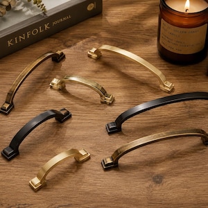 May include: A collection of seven arched cabinet pulls in various finishes including matte black, brushed gold, and antique brass. Each handle features a curved bar design with square mounting bases at both ends, arranged in a scattered layout.