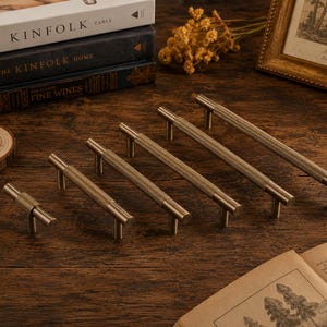 May include: A set of five brass-toned metal cabinet pulls arranged in a row by size, each featuring a cylindrical bar with a textured, knurled surface and two vertical mounting posts. The hardware has a metallic finish and a uniform design.