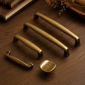 Può includere: Una collezione di quattro maniglie per armadietti in ottone anticato di varie lunghezze e un pomello quadrato. Ogni pezzo presenta una finitura metallica testurizzata e invecchiata con un design leggermente curvo e piedini di fissaggio.