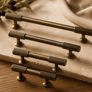 May include: Four brass cabinet pulls of varying lengths arranged in a descending stack. Each pull features a smooth central rod flanked by two cylindrical ends with a vertical ribbed texture and two circular mounting bases for attachment.