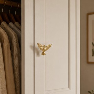 Puede incluir: Un pomo de armario de metal dorado con forma de p&aacute;jaro con las alas extendidas, montado en una puerta de panel beige claro. La figura del p&aacute;jaro presenta texturas de plumas detalladas en las alas y la cola, sirviendo como tirador funcional.