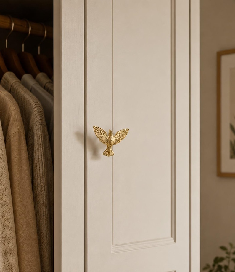 Puede incluir: Un pomo de armario de metal dorado con forma de p&aacute;jaro con las alas extendidas, montado en una puerta de panel beige claro. La figura del p&aacute;jaro presenta texturas de plumas detalladas en las alas y la cola, sirviendo como tirador funcional.