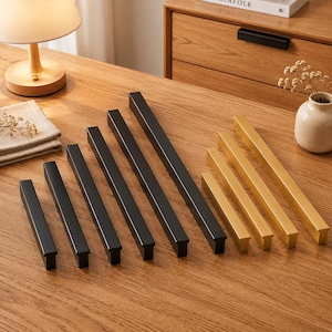 Puede incluir: Una colección de tiradores lineales para armarios dispuestos en fila, que incluye cinco tiradores de metal negro mate de varias longitudes y cuatro tiradores de metal dorado cepillado de varias longitudes, todos con perfil rectangular.