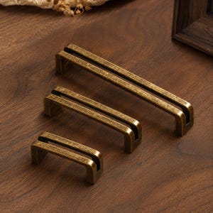 May include: Three rectangular cabinet pulls in varying lengths, each featuring a distressed antique brass finish with a textured surface. The handles have a U-shaped profile with a central open slot running along the top length of each piece.