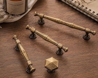 Puede incluir: Un juego de tres tiradores de armario de metal en tono latón antiguo y un pomo hexagonal a juego. Cada tirador presenta un diseño geométrico central facetado con dos puntos de montaje y un acabado texturizado y desgastado en la superficie.