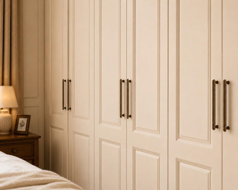May include: A set of tall, off-white wardrobe doors featuring recessed rectangular panels and vertical molding. Each door is equipped with a pair of long, slender, dark bronze-toned metal bar handles mounted vertically for easy access.