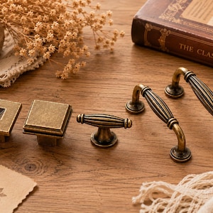 May include: A collection of antique brass hardware including two square knobs with beveled edges, one small fluted T-shaped knob, and two arched drawer pulls with vertical fluted detailing, all featuring a weathered, dark metallic finish.