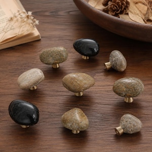 May include: A collection of eight cabinet knobs featuring smooth, rounded river stones in various shades of gray, black, and tan, each mounted on a small, circular brass-toned metal base. The stones vary in size and natural texture.