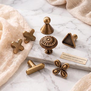 May include: A collection of six brass cabinet knobs in various shapes including a round floral knob, a triangular knob, a T-bar, two cross-shaped knobs, and a conical knob. A small rectangular card reads VINTAGE BRASS HARDWARE COLLECTION.