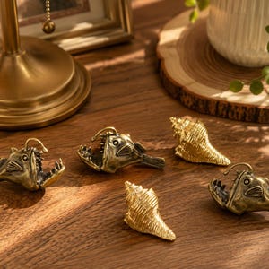 May include: A collection of five small metal figurines consisting of three brass-toned anglerfish with open mouths and prominent lures, alongside two textured gold-toned seashell ornaments arranged on a wooden surface.