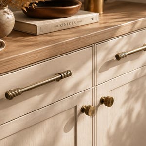 May include: Two long, horizontal, cylindrical brass cabinet pulls with ribbed detailing on the ends, and two matching round, ribbed brass cabinet knobs installed on light-colored wooden cabinet doors with a smooth, matte finish.