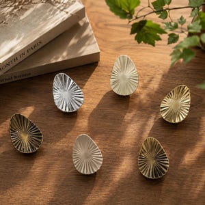 May include: Six teardrop-shaped cabinet knobs arranged in a circle, featuring a sunburst pattern with radiating lines. The set includes metallic finishes in silver, gold, and bronze, as well as a matte white finish, each with a textured surface.