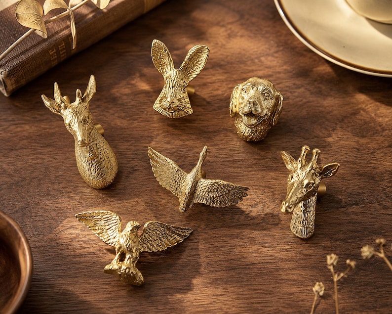 Puede incluir: Una colecci&oacute;n de seis pomos de armario de metal dorado con forma de cabezas y figuras de animales, incluyendo un ciervo, un conejo, un perro, una jirafa y dos aves con las alas extendidas. Cada pomo presenta texturas detalladas.