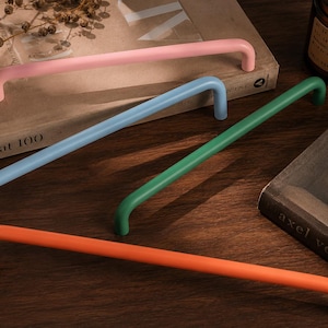 May include: Four long, tubular cabinet handles in matte finishes of pastel pink, light blue, forest green, and bright orange. Each handle features a smooth, curved design with two mounting points, presented as a set of hardware for furniture.
