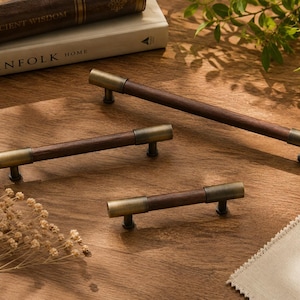 May include: Three cabinet pulls featuring a cylindrical dark brown wooden centre bar and brushed brass metal end caps. The hardware pieces are arranged in a descending size order, showcasing a combination of natural wood and metallic finishes.