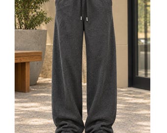 Könnte beinhalten: Dunkelgraue Jogginghose mit weitem Bein, elastischem Bund mit schwarzem Kordelzug und zwei Seitentaschen. Die Hose besteht aus einem weichen, strukturierten Stoff und wird mit weißen Sneakern kombiniert.