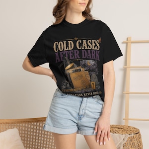 May include: A black short-sleeve t-shirt featuring the text COLD CASES AFTER DARK in tan and purple. The graphic includes a desk lamp, a file folder labeled CASE FILE, a fingerprint, a number 7 evidence marker, and SOME FILES NEVER SLEEP.
