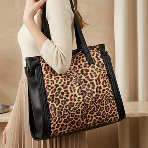 May include: A large tote bag featuring a central panel with a brown and black leopard print pattern, flanked by solid black side panels and black shoulder straps. The bag has a rectangular shape with visible stitching and two front pockets.
