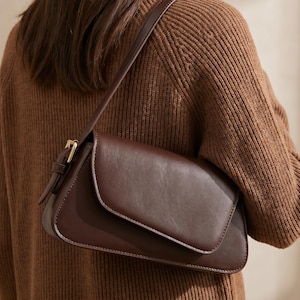 May include: A dark brown shoulder bag with a smooth finish, featuring a fold-over flap closure, a single adjustable strap with a gold-toned metal buckle, and rounded edges, worn over a brown ribbed knit sweater.