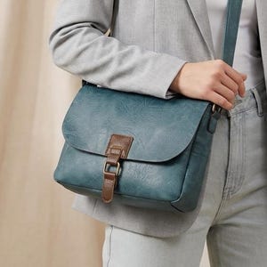 May include: A teal blue crossbody bag with a rectangular flap, featuring a brown leather-like strap closure with a metal buckle. The bag has a matching teal blue shoulder strap and a smooth, matte finish, worn over a light gray blazer and white top.