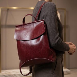 Peut inclure: Un sac à dos en cuir bordeaux profond présentant une silhouette lisse et structurée avec un grand rabat repliable, une poignée de transport supérieure et des bretelles réglables. Le matériau a un éclat subtil et une esthétique épurée.