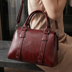 May include: A deep burgundy handbag with a smooth, polished finish, featuring two top handles and a detachable shoulder strap. The bag includes vertical decorative straps on the front, secured with small metal rivets and silver-toned hardware rings.