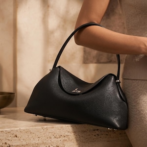 May include: A black leather handbag with a pebbled texture, featuring a single arched handle, a fold-over flap closure with a small silver-toned metallic accent, and a structured base with visible silver-toned feet for stability.
