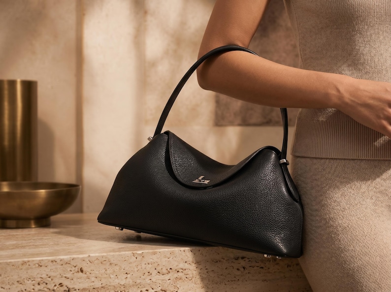 May include: A black leather handbag with a pebbled texture, featuring a single arched handle, a fold-over flap closure with a small silver-toned metallic accent, and a structured base with visible silver-toned feet for stability.