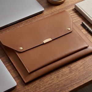 May include: A tan document sleeve with a flap closure, featuring two gold-toned metal snap buttons and a central gold-toned metal rectangular accent piece. The sleeve has visible stitching along the edges and a smooth, uniform surface texture.