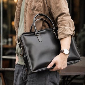 May include: A black leather briefcase featuring a rectangular silhouette, two top handles, and a side zip closure. The bag has a smooth, polished finish with visible stitching along the edges and metallic hardware attachments for a shoulder strap.