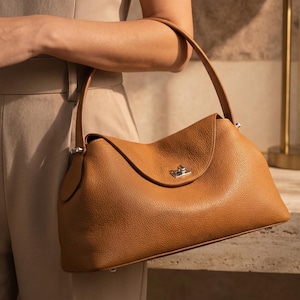 May include: A tan leather handbag with a pebbled texture, featuring a single shoulder strap, a curved top flap, and a silver-toned metal turn-lock closure. The bag has a structured base with small metal feet and silver hardware attachments.