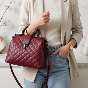 May include: A deep burgundy doctor-style handbag featuring a quilted diamond pattern, a structured frame, and a central metal clasp closure. The bag includes a top handle and a matching adjustable shoulder strap with dark metal hardware accents.