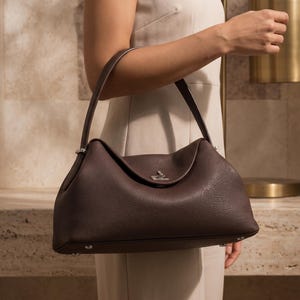 May include: A dark brown leather handbag with a pebbled texture, featuring a single top handle, a curved silhouette, and a silver-toned metal turn-lock closure at the center. The bag includes silver-toned hardware accents on the handle attachments.