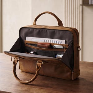 May include: A tan leather briefcase with a top handle and a zipped main compartment. The interior is lined with dark fabric and features multiple pockets containing a document, a small wallet, a smartphone, and other small personal items.