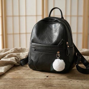 May include: A black faux leather backpack featuring a top handle, adjustable shoulder straps, and a front zippered pocket. The bag is accented with a white fluffy pom-pom keychain attached to the zipper pull and includes side pockets.